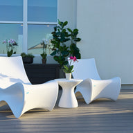Two modern white chairs and a Ledge Lounger Autograph Standard Side Table with a potted orchid are arranged on a wooden patio deck, surrounded by green plants and large windows in the background.