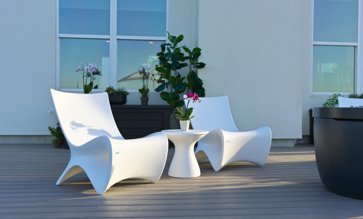 Two modern white chairs and a Ledge Lounger Autograph Standard Side Table with a potted orchid are arranged on a wooden patio deck, surrounded by green plants and large windows in the background.