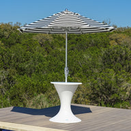 A black and white striped umbrella rests in the Ledge Lounger Autograph Umbrella Stand Ice Bin on a wooden deck by a pool, framed by green trees and a clear blue sky.