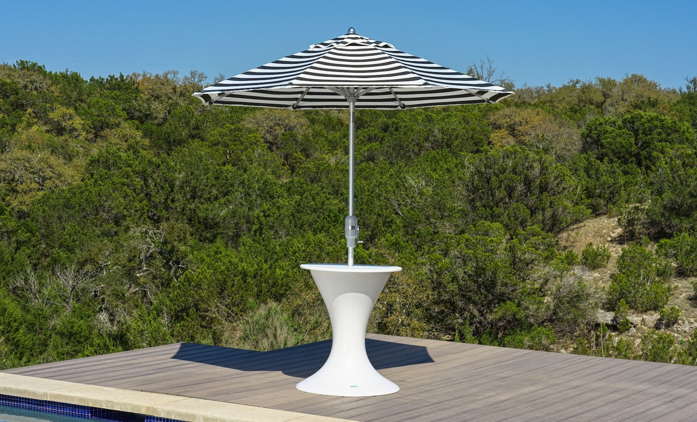 A black and white striped umbrella rests in the Ledge Lounger Autograph Umbrella Stand Ice Bin on a wooden deck by a pool, framed by green trees and a clear blue sky.
