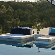 Two Ledge Lounger Affinity Square Sunbeds with flat cushions and blue and white pillows sit beside an infinity pool, paired with a potted orchid and large umbrella, overlooking a lush forest—stylish outdoor relaxation.