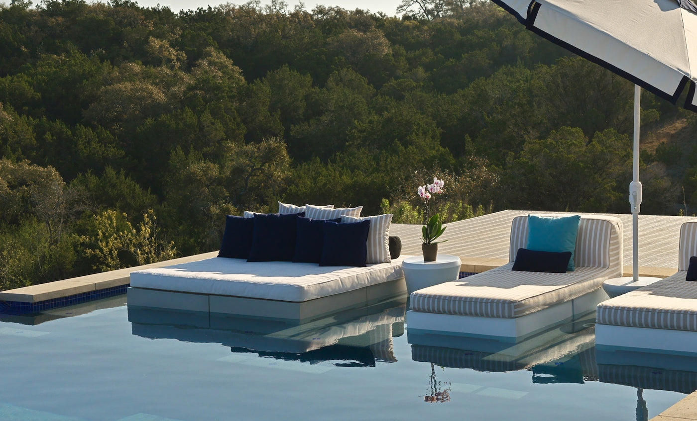 Two Ledge Lounger Affinity Square Sunbeds with flat cushions and blue and white pillows sit beside an infinity pool, paired with a potted orchid and large umbrella, overlooking a lush forest—stylish outdoor relaxation.
