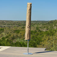 A Ledge Lounger Umbrella Cover in beige protects a closed umbrella on a wooden deck by the pool, framed by green hills and a clear blue sky.