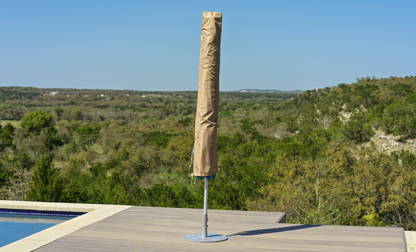 A Ledge Lounger Umbrella Cover in beige protects a closed umbrella on a wooden deck by the pool, framed by green hills and a clear blue sky.