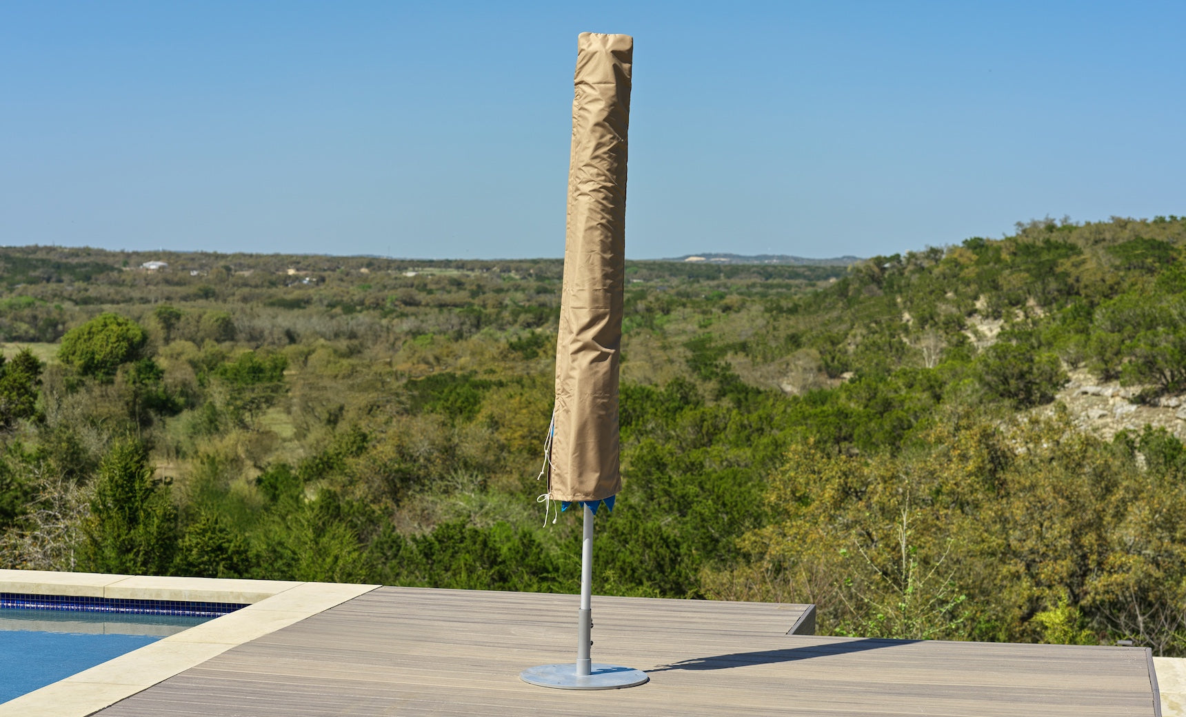 A Ledge Lounger Umbrella Cover in beige protects a closed umbrella on a wooden deck by the pool, framed by green hills and a clear blue sky.