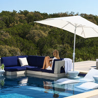 A woman relaxes on the blue Ledge Lounger Signature Sectional Middle with two-piece cushions beneath a large white umbrella by a modern pool, surrounded by loungers and lush greenery.