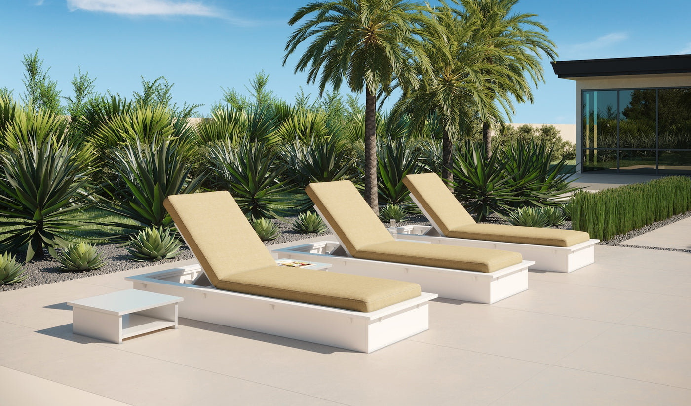 Three Echo Chaise loungers from Ledge Lounger, featuring white frames and beige fabric, are set on a tiled patio amidst lush palm trees. A small side table complements this tranquil outdoor setting under a clear blue sky with a modern building on the right.