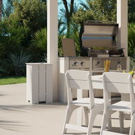 Outdoor dining is a delight with a white table and Ledge Loungers Mainstay Dining Side Chairs, perfectly set near a stainless steel grill. Plates, utensils, glasses, and vases adorn the scene amidst lush green foliage under a clear blue sky.