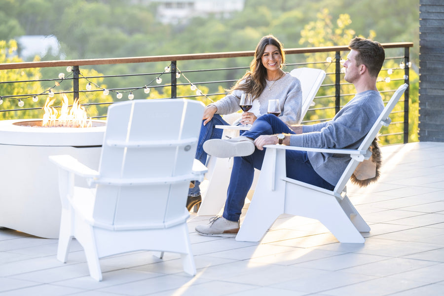 A woman and a man relax on Mainstay Ultimate Adirondack chairs by Ledge Lounger on the patio, sipping wine. A fire pit flickers between them, with lush greenery and string lights in the background, creating a serene atmosphere.