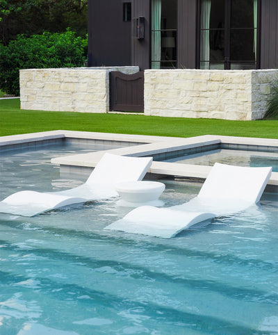 Two white in-pool chaise loungers with a side table on a baja shelf of a backyard pool.