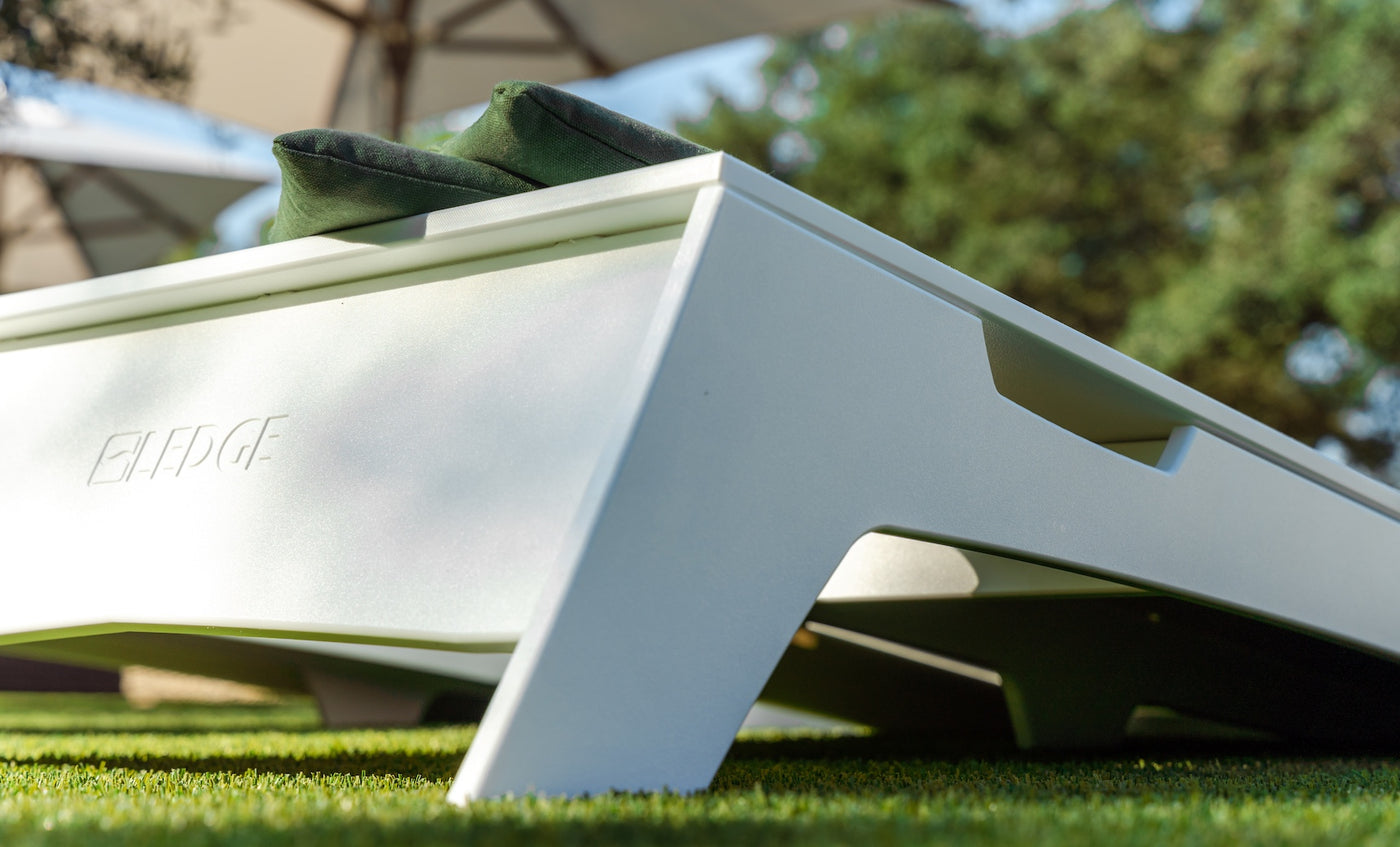 Close-up of a modern white outdoor lounge chair with two green cushions, set on artificial grass beside a Ledge Lounger Cornhole set. Blurred trees and parasols are in the background; ELLEFGE is engraved on the side.