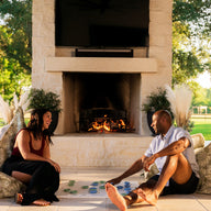 A woman and a man smile and play the Ledge Lounger Oversized Patio Games Set on large cushions by an outdoor fireplace, enjoying a sunny patio surrounded by green trees and a wooden fence.