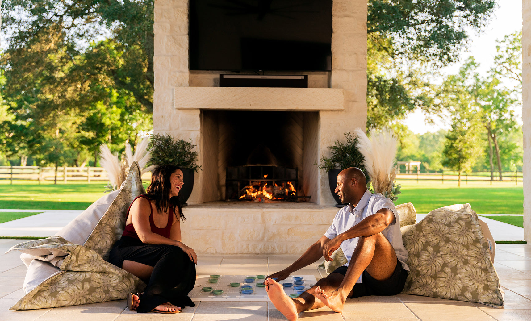 A woman and a man smile and play the Ledge Lounger Oversized Patio Games Set on large cushions by an outdoor fireplace, enjoying a sunny patio surrounded by green trees and a wooden fence.