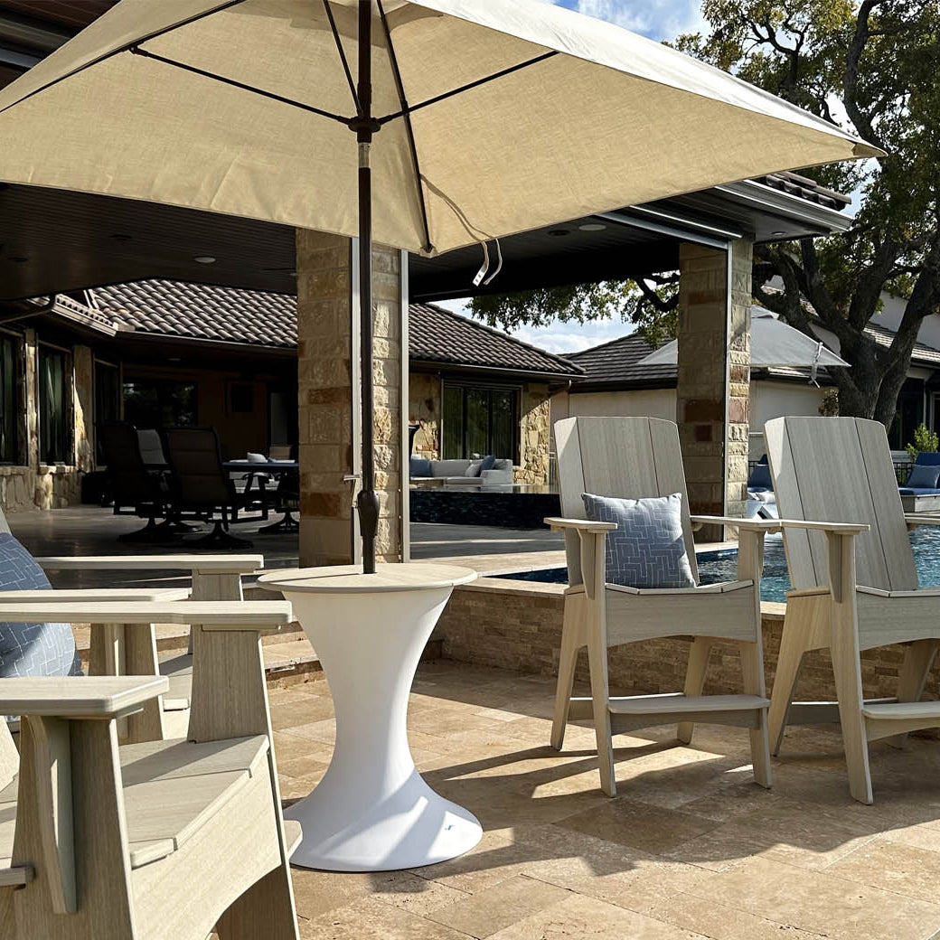 A patio featuring wooden chairs and a small white table is shaded by the Essential Fiberglass Umbrella - 11 Octagon Pulley from Ledge Lounger. This wind-resistant, large beige umbrella is set by a clear pool on a stone-paved deck, with a modern house and trees in the background.