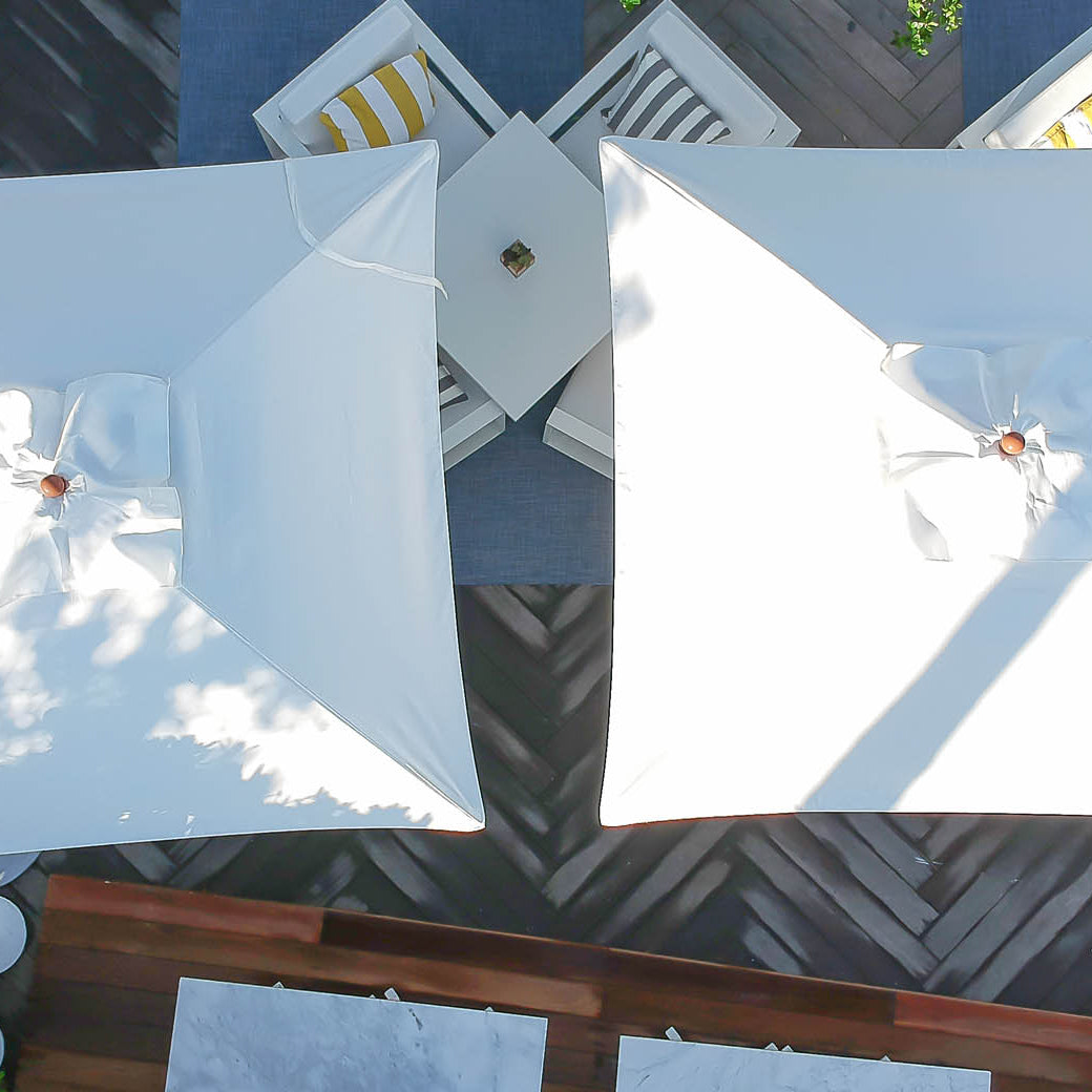 Aerial view of two Ledge Lounger Essential Fiberglass Umbrellas casting shadows over outdoor furniture, including a table with chairs featuring yellow and white striped cushions. The sunny scene, surrounded by greenery, highlights the durable marine-grade fabrics.