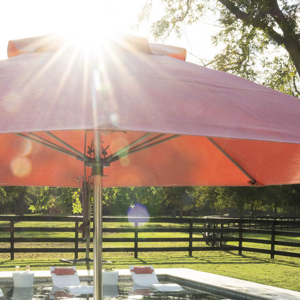 The Essential Fiberglass Umbrella - 6.5 Square Crank Auto Tilt by Ledge Lounger shades a pool with white lounge chairs, as sunlight filters through trees over a fenced grassy area, with durable marine-grade fabrics enhancing its resilience against the elements.