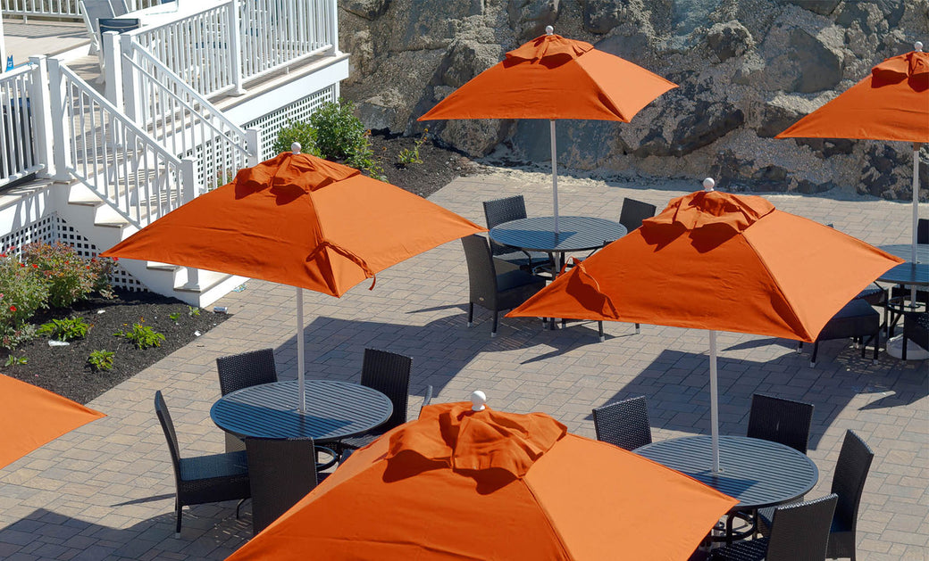 An outdoor seating area with round tables and chairs beneath large Ledge Lounger Essential Fiberglass Umbrellas - 7.5 Square Pulley on a paved patio. A white railing and stairs are visible on the left, against a rocky backdrop, combining style and durability with marine-grade fabrics.