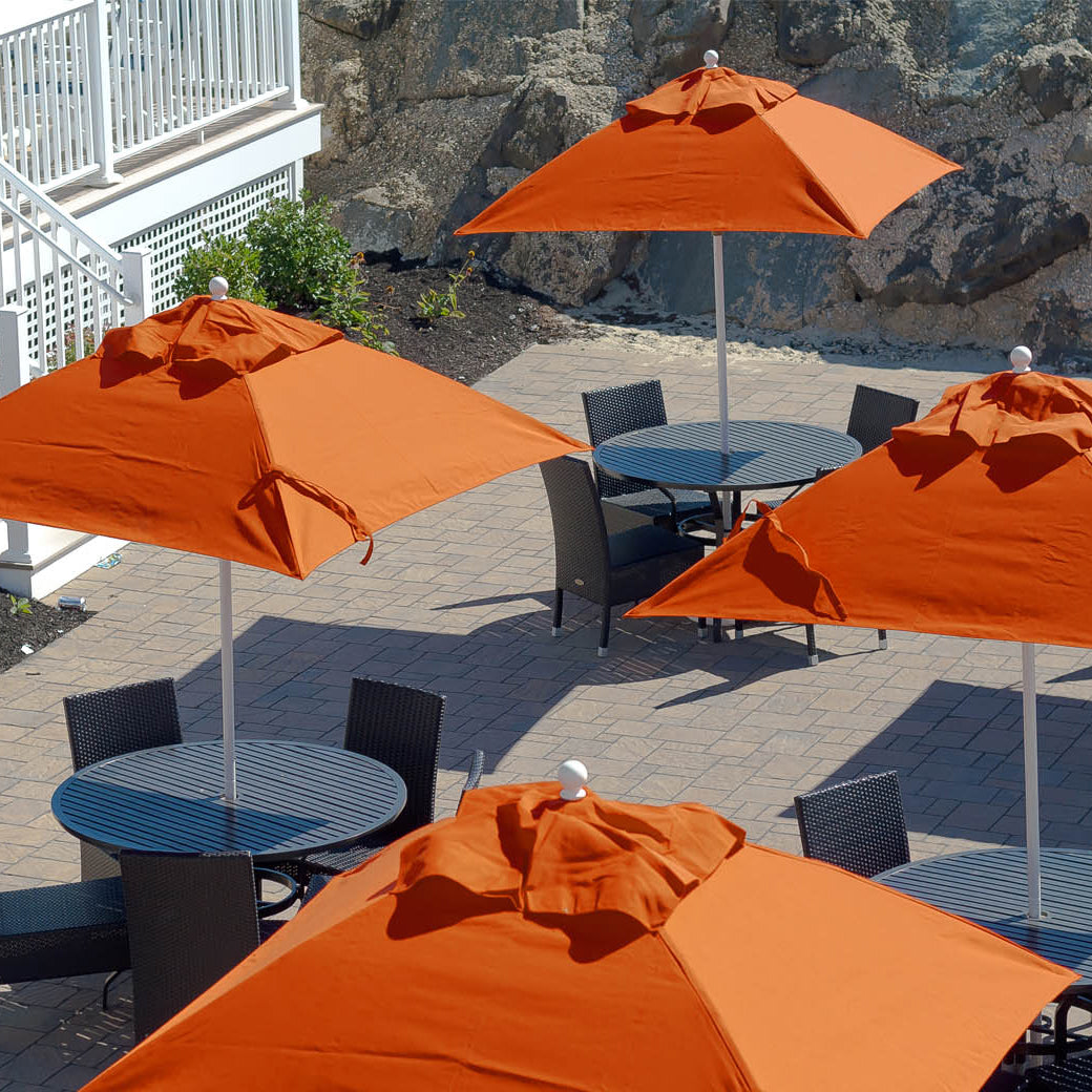 The outdoor patio features Ledge Loungers Essential Fiberglass Umbrella - 6.5 Square Crank Auto Tilt shading round tables and black chairs. The area is paved with light gray bricks, bordered by rocky landscaping, and has a white corrosion-resistant railing on the left.