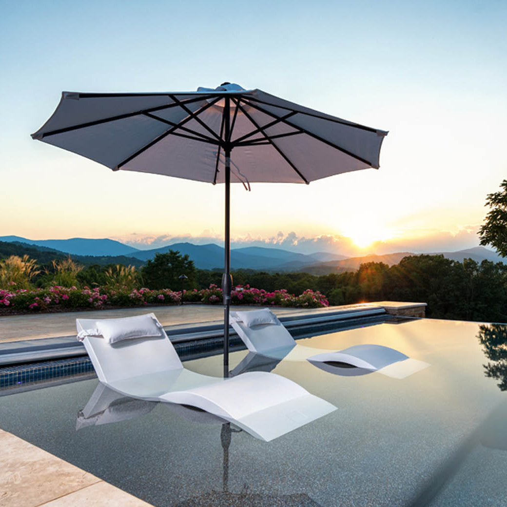 A serene infinity pool with two submerged lounge chairs featuring marine-grade fabrics and a Ledge Lounger Essential Fiberglass Umbrella - 11 Octagon Pulley overlooks a scenic mountain view at sunset, the clear sky highlighting lush greenery and vibrant flowers.
