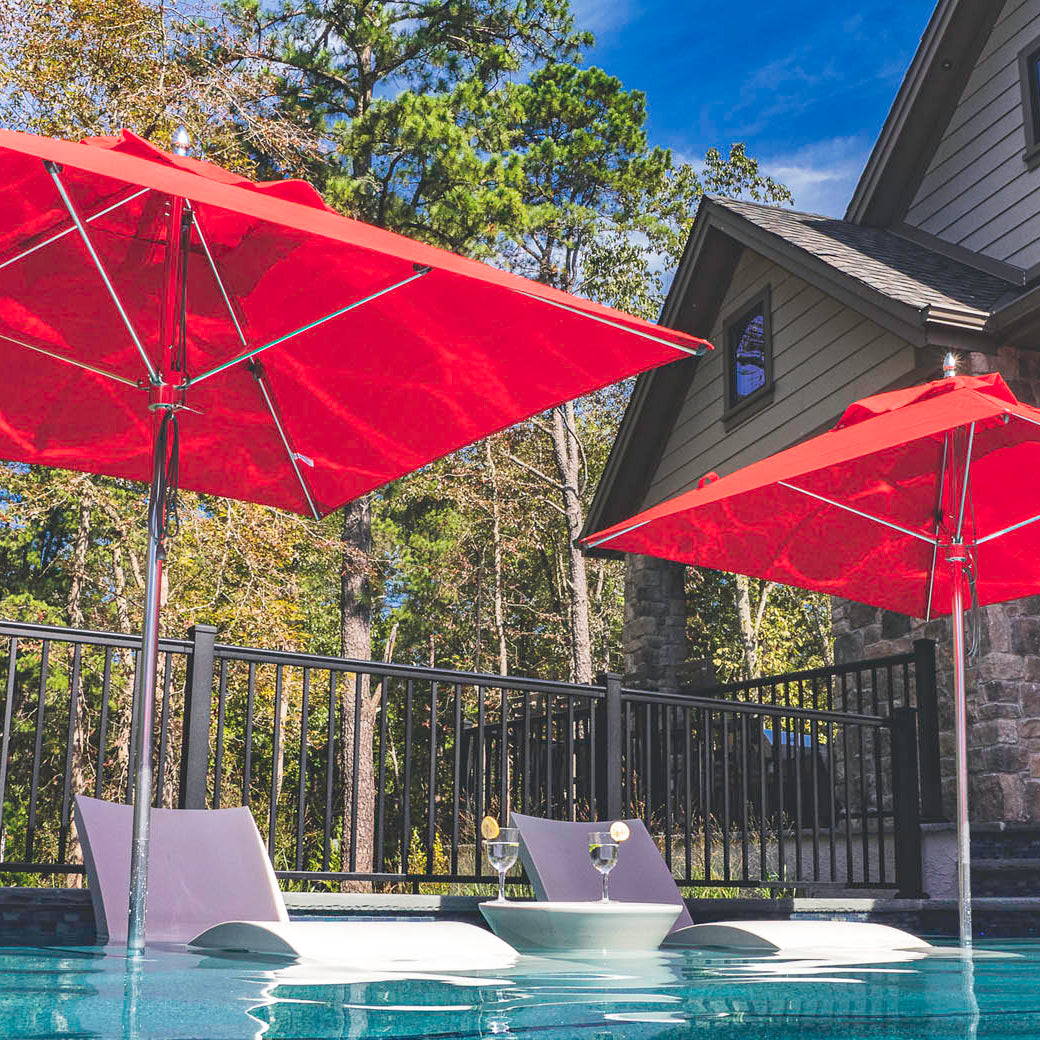 Two Premier Aluminum Umbrella - 9 Octagon Pulley, in red by Ledge Lounger, shade white lounge chairs by a stone-walled house pool. Trees encircle this serene oasis under a clear blue sky.