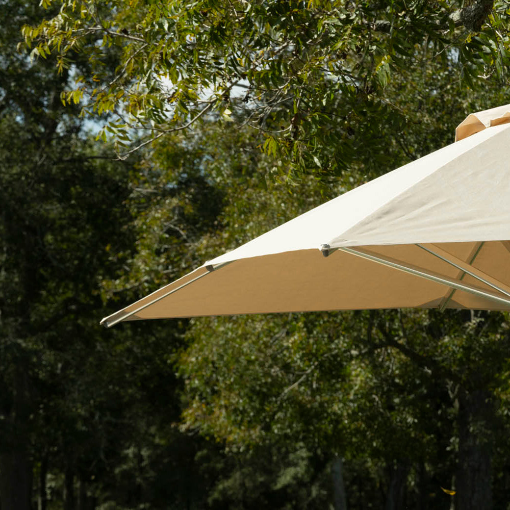 The Ledge Lounger Premier Aluminum Umbrella - 6.5 Square Pulley, in beige, partially opens against lush green trees. Its marine-grade fabrics and wind-resistant design interact with sunlight, suggesting a sunny day in a wooded setting.