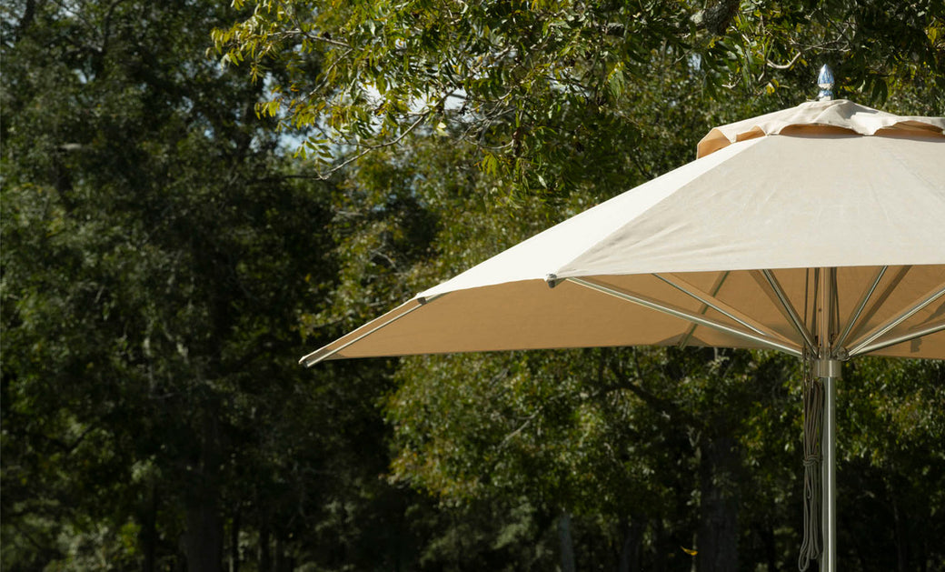 A Premier Aluminum Umbrella - 9 Octagon Pulley by Ledge Lounger offers shade beneath lush trees on a sunny day. Made with marine-grade fabrics, the partially open large beige umbrella combines style and wind resistance against the backdrop of dense foliage and dappled sunlight.