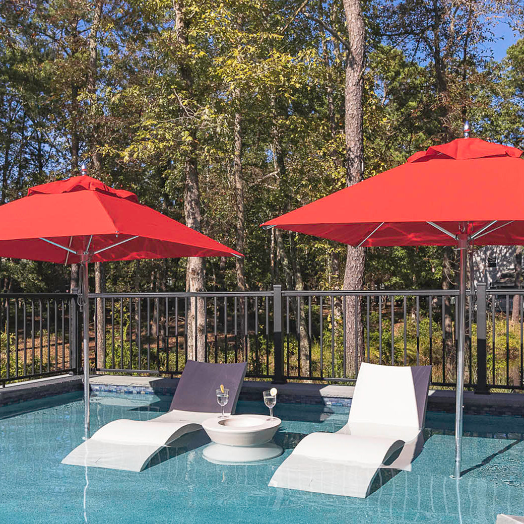 Two modern lounge chairs with a small round table and glasses sit in a shallow pool. Two Ledge Lounger Premier Aluminum Umbrellas - 9 Octagon Pulley, crafted with marine-grade fabrics, offer shade. A black metal fence and tall trees surround the area, creating a serene outdoor setting of tranquility.