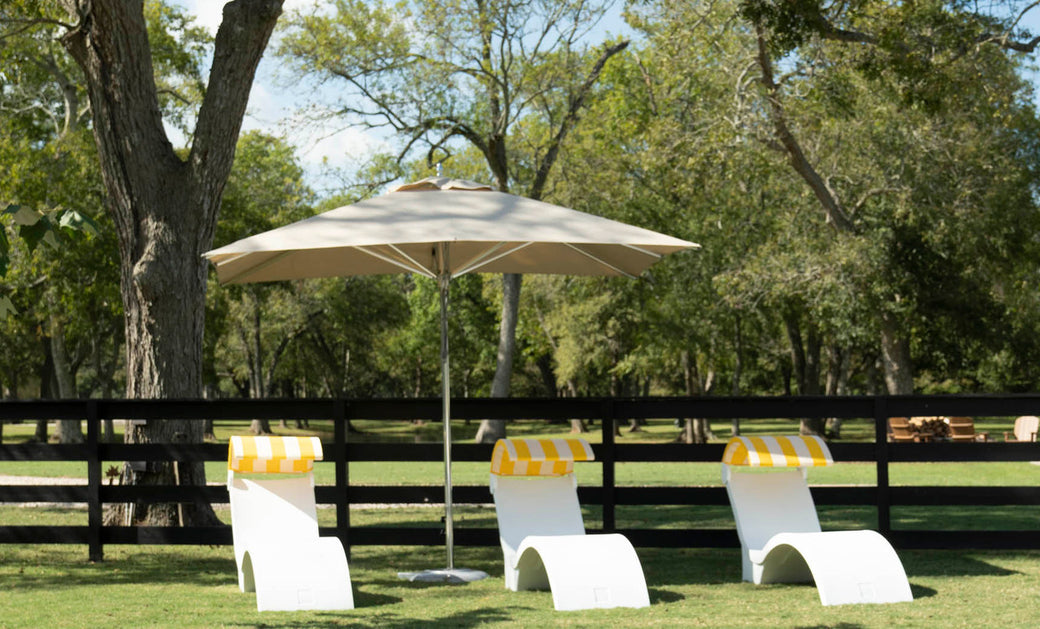 Three white lounge chairs with yellow and white striped towels sit under a Ledge Lounger Premier Aluminum Umbrella - 9 Octagon Pulley on the grassy lawn, with tall trees and a black wooden fence visible against the clear blue sky.
