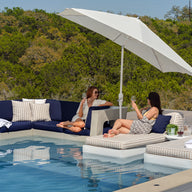 Two women relax on a luxurious patio set by the pool, enjoying resort-level luxury. They sit beneath a Ledge Lounger 7.5 Octagon Signature 360° Tilt Umbrella, surrounded by trees. The area features a navy couch and striped loungers, with a towel and drinks enhancing the serene scene.
