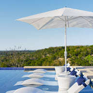 A tranquil outdoor scene showcases Ledge Loungers rustproof white lounge chairs with blue cushions, partially submerged in an infinity pool. A 6.5 Square Signature 360° Tilt Umbrella offers shade, set against a vast tree-covered landscape under a clear blue sky.