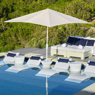 A modern poolside oasis includes white lounge chairs partially submerged in water, perfectly shaded by a Ledge Lounger 7.5 Octagon Signature 360° Tilt Umbrella. A double daybed with blue and white cushions sits on a wooden deck amidst lush greenery.