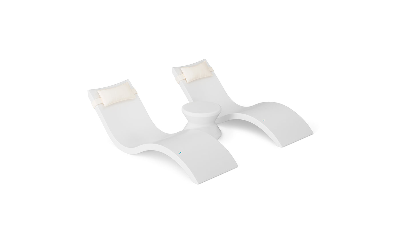 The Ledge Lounger Signature Chaise Headrest Pillow and Side Table In-Pool Set offers two modern white loungers with cream headrest pillows, connected by a round table—perfect for poolside relaxation against a sleek white backdrop.