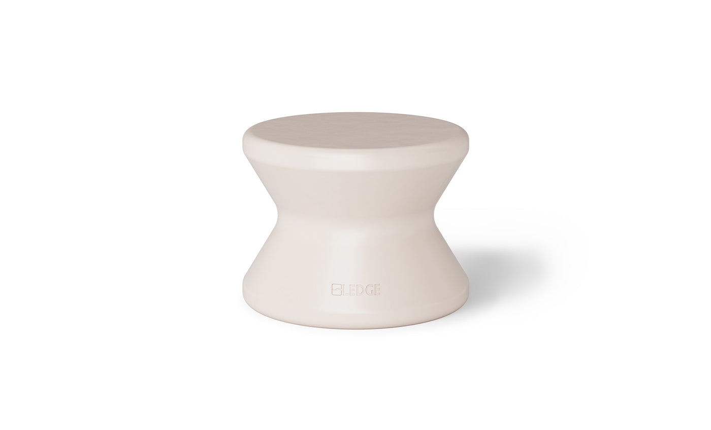 The Signature Standard Side Table by Ledge Lounger is a small, hourglass-shaped, light beige stool with a smooth surface for in-pool use on sun shelves. It features the word EDGE inscribed near the bottom, adding elegance and functionality.