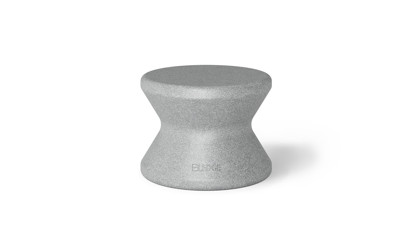 The Signature Standard Side Table by Ledge Lounger is a gray hourglass-shaped stool with a textured surface and smooth, curved design, perfect for in-pool use. The name BLØK is subtly engraved near the base, set against a plain white background.