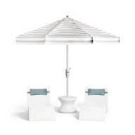 Two white lounge chairs with blue cushions flank a small white table, all shaded by the Ledge Lounger 7.5' Octagon Signature 360° Tilt Umbrella in classic white and gray stripes.