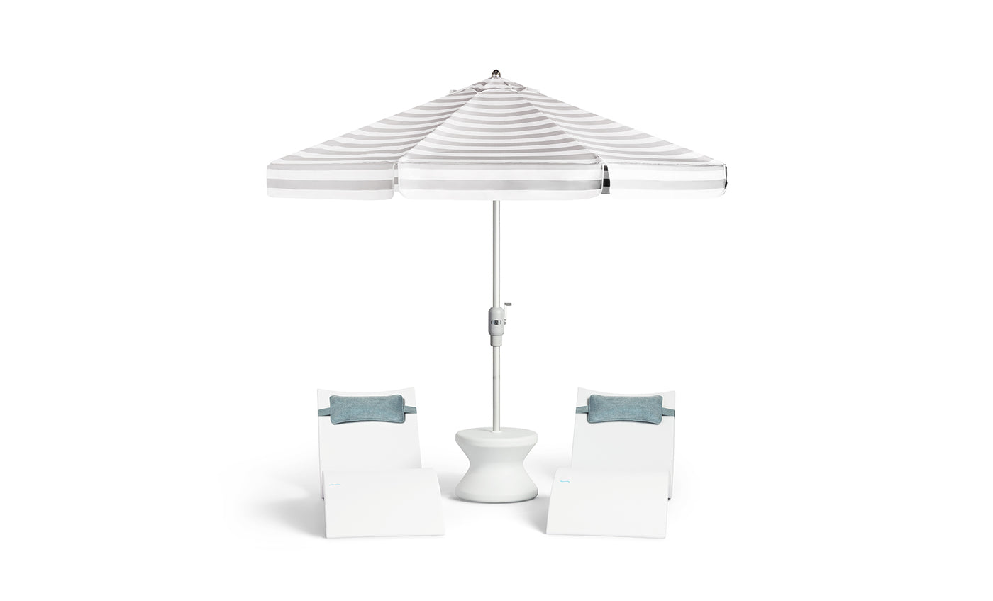 Two white lounge chairs with blue cushions flank a small white table, all shaded by the Ledge Lounger 7.5' Octagon Signature 360° Tilt Umbrella in classic white and gray stripes.