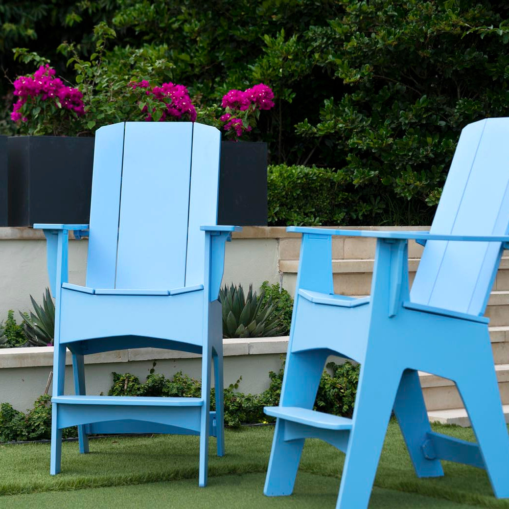 Two oversized Mainstay Adirondack Tall chairs by Ledge Lounger in light blue mark the ideal outdoor lounge setting. Set on the grass, concrete steps ascend behind with pink flowers flourishing. The vibrant garden scene is enhanced by built-in bottle openers for added convenience.