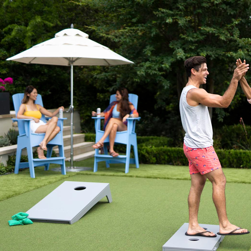 Two men exchange a high-five on the lawn near cornhole boards, while two women engage in conversation, relaxing in Ledge Loungers Mainstay Adirondack Tall chairs under a large umbrella. Theyre dressed in summer attire amid greenery, with a lounge seat featuring a built-in bottle opener nearby.