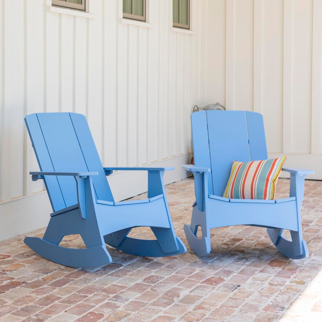 Two light blue Mainstay Adirondack Rockers by Ledge Lounger rest on a brick patio beside a wooden door, enhancing the serene space. One chair has a striped cushion. The white paneled wall with three small windows elegantly complements the setting.