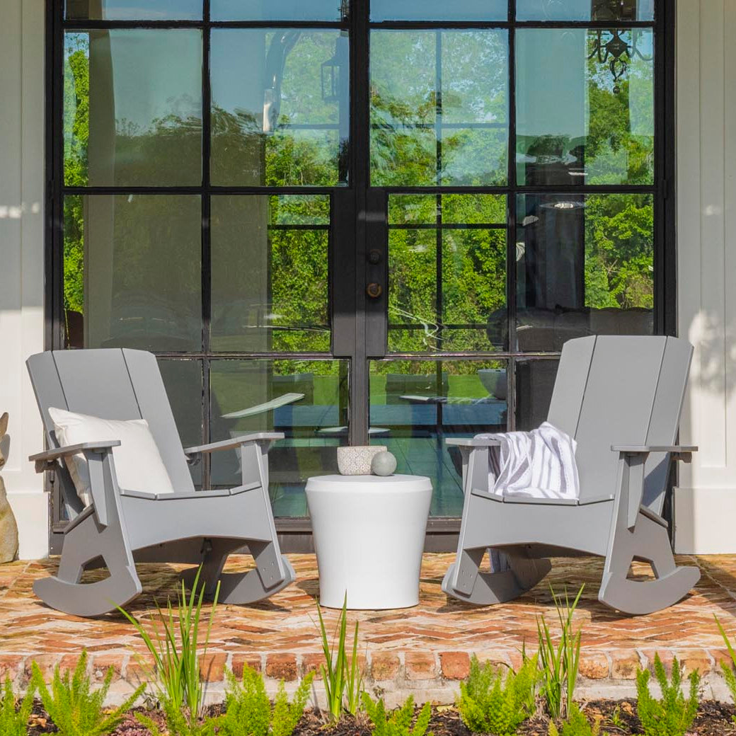 Two Mainstay Adirondack Rockers by Ledge Lounger, in gray, with a white side table between them sit on a brick patio. A striped blanket on one chair adds coziness. Large glass doors with lush greenery visible through the panes are in the background.