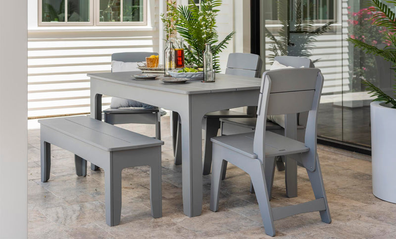 An outdoor dining area showcases the Ledge Lounger Mainstay Rectangular Dining Table in sleek gray, complemented by matching chairs and a bench. The table is adorned with glass bottles, a vase plant, and tableware on a stone-tiled patio adjacent to a glass door with white siding and lush potted plants.