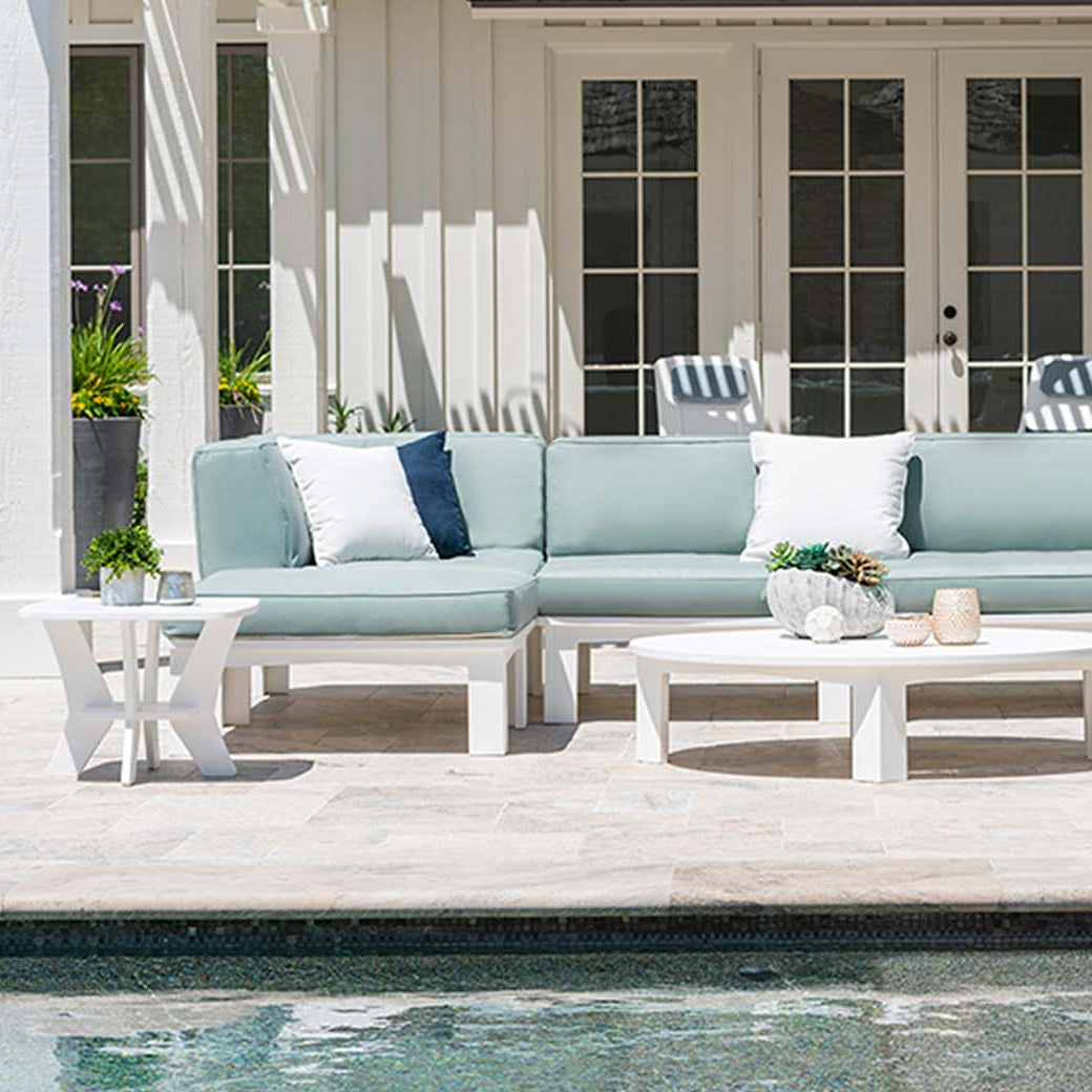 In a patio setting by the pool, the Mainstay Sectional Corner by Ledge Lounger is arranged in white and blue hues around white coffee tables. Tall planters add greenery, while a white building with large windows sets the backdrop, offering limitless arrangement options.