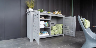 A modern outdoor storage cabinet with open doors, revealing folded towels, green and white patterned cushions, bottled drinks, and glasses. Small plants and more bottles are on top. It is set against a dark wall on a tiled floor.