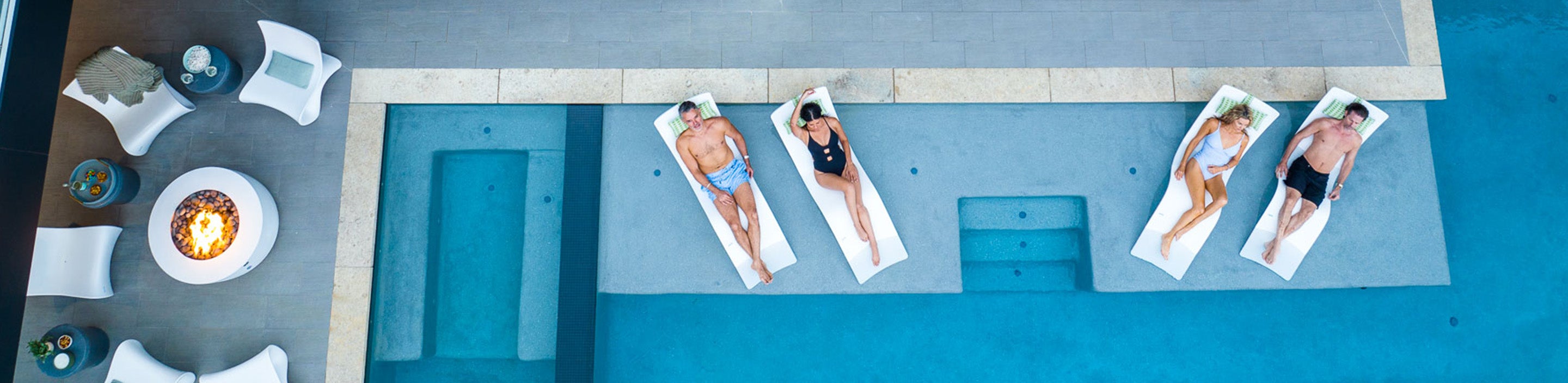 Four people are sunbathing on lounge chairs partially submerged in a pool, surrounded by a patio with white chairs and a lit fire pit. The scene is viewed from above.