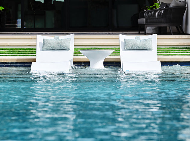Two white lounge chairs partially submerged in a swimming pool face a circular white side table. Steps lead to a patio with a dark couch and potted plants in the background, creating a serene outdoor setting.