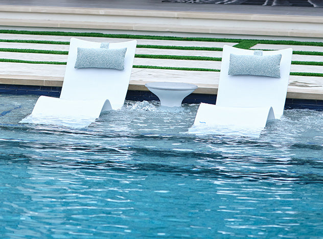 Two modern white lounge chairs with light gray pillows are positioned in a shallow pool area. A small white round table is placed between them. The water is clear, and a grassy area is visible in the background.