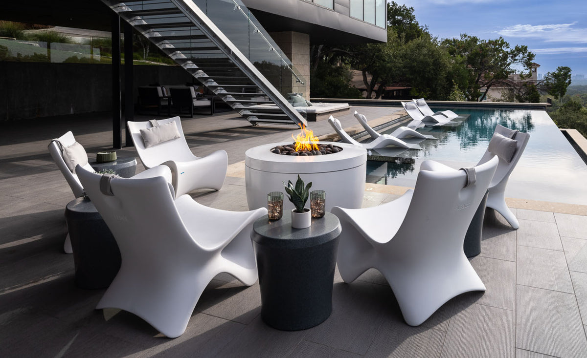 Modern outdoor patio with white sculpted chairs around a fire pit, small side tables with plants and candles, next to a pool with partially submerged loungers, and a glass staircase by a contemporary building.