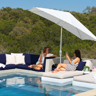 Two women relax and chat on modern poolside furniture, featuring Ledge Loungers Autograph Standard Side Table, cushioned loungers, and a sectional sofa under a large white umbrella, with trees and blue sky in the background.