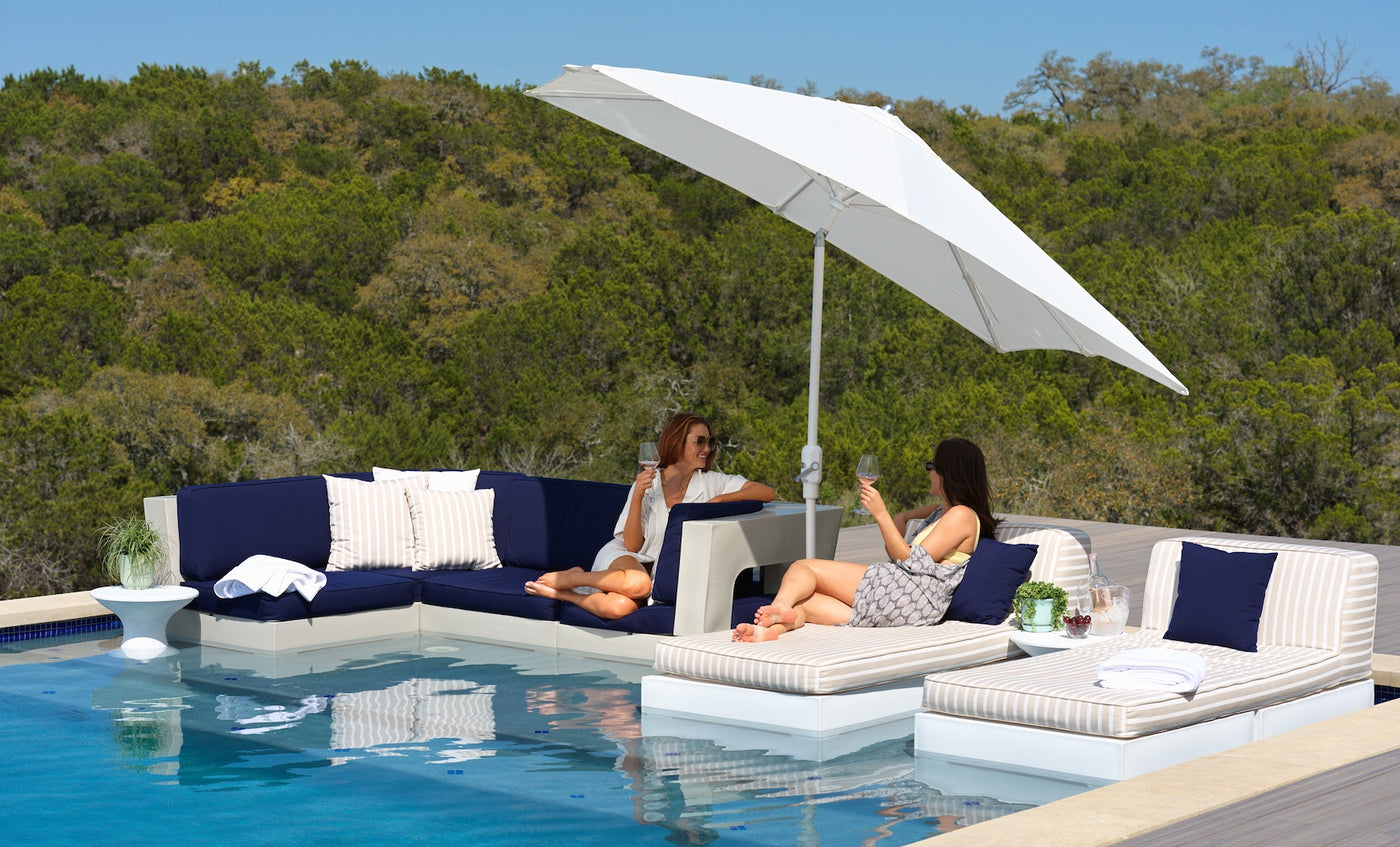 Two women relax and chat on modern poolside furniture, featuring Ledge Loungers Autograph Standard Side Table, cushioned loungers, and a sectional sofa under a large white umbrella, with trees and blue sky in the background.