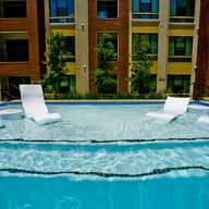 The Ledge Lounger Signature Chaise with Chairs and Side Table In-Pool 6-Piece Set sits partially submerged in a clear blue pool, with a modern apartment building and green trees visible in the background under bright sunlight.